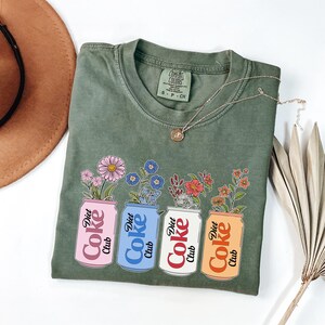 May include: Olive green t-shirt with a floral design. The graphic features four Diet Coke cans in pink, blue, white, and orange, each holding a different type of flower. A silver necklace and a brown hat are also visible.