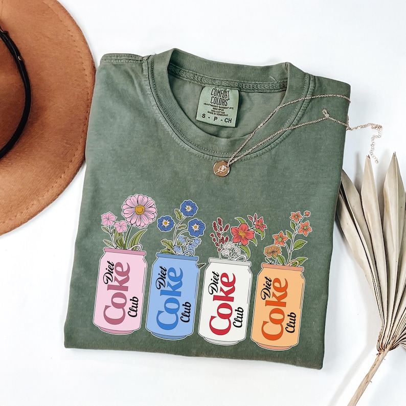 May include: Olive green t-shirt with a floral design. The graphic features four Diet Coke cans in pink, blue, white, and orange, each holding a different type of flower. A silver necklace and a brown hat are also visible.