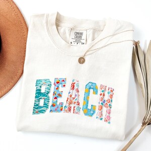 Puede incluir: Una camiseta de manga corta color crema con la palabra BEACH en grandes letras de tela aplicadas. Cada letra muestra un patrón único, incluyendo olas del mar, pelotas de playa, coral, soles y conchas. Un collar de tono dorado descansa sobre el cuello.