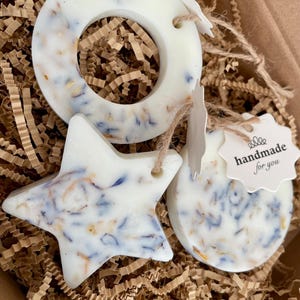 May include: A gift box containing three white wax melts shaped like a star, a circle, and a crescent moon. The wax melts are infused with dried flowers and have a tag that says "handmade for you". The box is filled with brown crinkle paper.