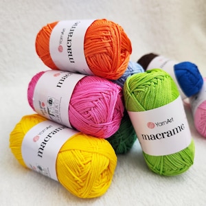 Puede incluir: Una colección de ovillos de cordón YarnArt Macrame en colores vibrantes que incluyen naranja, rosa brillante, amarillo, verde lima, azul y marrón oscuro. Cada ovillo tiene una etiqueta de papel blanco con la marca YarnArt y la palabra macrame.
