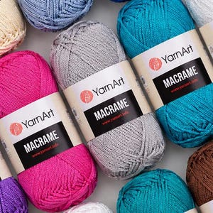 Könnte beinhalten: Eine Sammlung von YarnArt Macrame Garnknäueln in verschiedenen Farben, darunter leuchtendes Pink, Silbergrau, Türkis, Blau, Lila, Weiß und Braun. Jedes Knäuel hat ein Papieretikett mit dem YarnArt-Logo und dem Text MACRAME und www.yarnart.info.