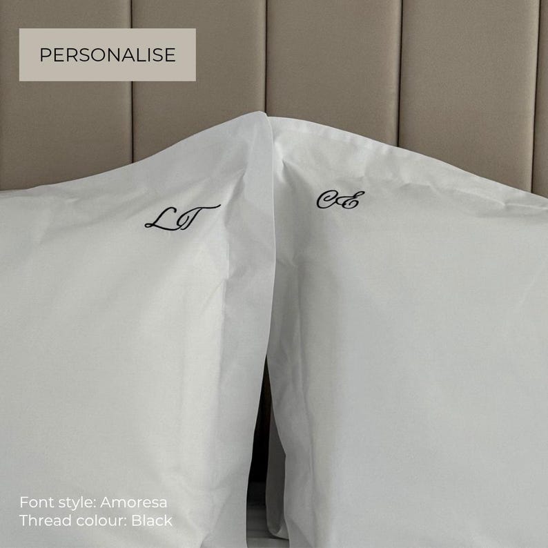 Op de afbeelding: Twee witte kussenslopen met zwarte geborduurde monograminitialen. De linker kussensloop toont de letters LT, terwijl de rechter kussensloop de letters CE bevat. De tekst PERSONALIZE, Font style: Amoresa en Thread colour: Black zijn zichtbaar.