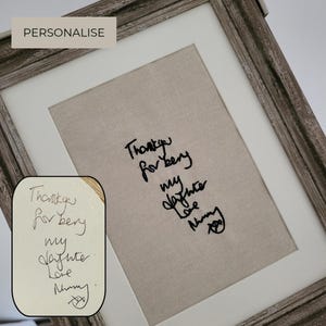 Può includere: Un quadro da parete con sfondo in tessuto beige e ricamo nero che riproduce una scritta a mano: Thank you for being my daughter Love Mummy. La cornice è realizzata in legno grigio effetto vissuto con un bordo bianco.