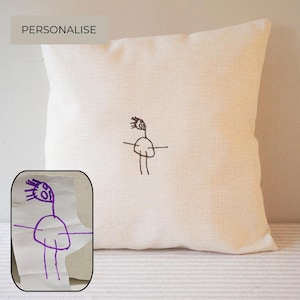 Puede incluir: Un cojín cuadrado de color blanco roto con un bordado negro centrado que muestra un dibujo lineal de una figura de palo con cuerpo redondo y pelo puntiagudo. Un recuadro muestra el dibujo original en tinta morada con el texto PERSONALIZE.