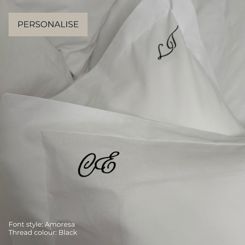Op de afbeelding: Witte kussenslopen met zwarte geborduurde monogrammen in het Amoresa-lettertype. De ene kussensloop toont de letters LT, de andere de letters CE. De tekst PERSONALISE is zichtbaar in een kader, met Font style: Amoresa en Thread colour: Black.