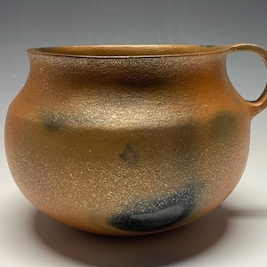 May include: A golden brown ceramic mug with a speckled glaze and a single handle. The mug has a rounded body and a slightly flared rim.