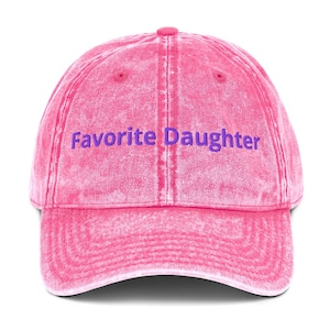 Op de afbeelding: Een roze baseballpet met de tekst "Favorite Daughter" in paars. De pet heeft een gebogen klep en een knoop aan de bovenkant. De stof heeft een licht vervaagde, vintage uitstraling.