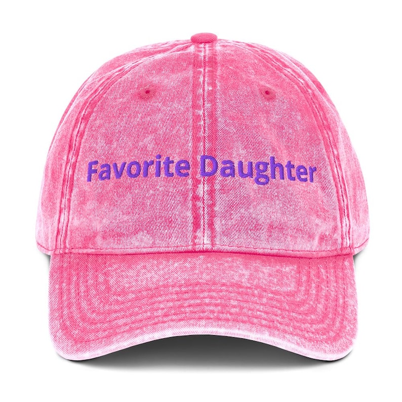 Op de afbeelding: Een roze baseballpet met de tekst "Favorite Daughter" in paars. De pet heeft een gebogen klep en een knoop aan de bovenkant. De stof heeft een licht vervaagde, vintage uitstraling.