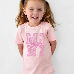 May include: A light pink t-shirt with the word "MINI" printed in a decorative font. The letters are a darker shade of pink and feature a floral pattern. A pink bow is between the letters. The shirt is worn by a young child.
