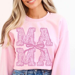 May include: A light pink sweatshirt with the word "MAMA" in large, floral-patterned letters. A pink bow connects the two "MA" letters. The sweatshirt is a soft, casual style.