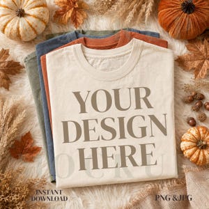 Könnte beinhalten: Ein Stapel aus vier gefalteten T-Shirts mit Rundhalsausschnitt in Creme, Rostrot, Blau und Olivgrün. Das oberste cremefarbene T-Shirt zeigt den Text "YOUR DESIGN HERE" in großen, fetten, braunen Großbuchstaben. Die Shirts liegen ordentlich.