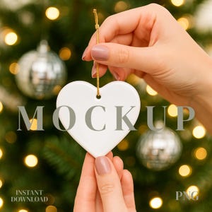 Può includere: Un ornamento bianco a forma di cuore con un filo d'oro, tenuto in mano. La parola "MOCKUP" è stampata sull'ornamento. Lo sfondo presenta un albero di Natale con ornamenti argentati e luci bokeh. Le parole "INSTANT DOWNLOAD" e "PNG" sono visibili.
