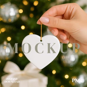 Könnte beinhalten: Ein weißer herzförmiger Christbaumschmuck mit dem Aufdruck "MOCKUP", der von einer Hand gehalten wird. Der Schmuck hängt an einem goldenen Faden. Der Hintergrund zeigt einen Weihnachtsbaum mit silbernen Ornamenten und Bokeh-Lichtern. Die Wörter "INSTANT DOWNLOAD" und "PNG" sind ebenfalls sichtbar.