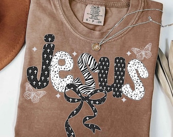 Puede incluir: Una camiseta marrón con la palabra "JESUS" en una fuente decorativa. Las letras tienen una mezcla de patrones, incluyendo cruces, rayas de cebra y lunares. También se presenta un lazo con cruces y dos mariposas. La camiseta tiene una etiqueta Comfort Colors.