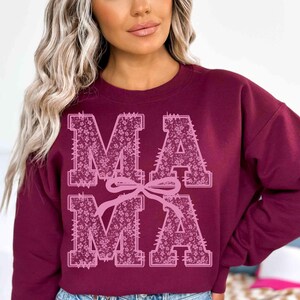 May include: Burgundy sweatshirt with the word "MAMA" in large, floral-patterned letters. The letters are in a block style with a pink bow connecting the two words. The sweatshirt is a solid burgundy color.
