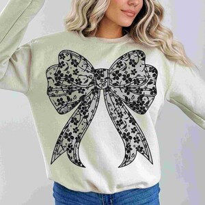 May include: A light beige sweatshirt with a large black lace bow design.