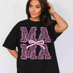 May include: Black t-shirt with the word "MAMA" in pink floral letters. A pink bow is centered below the letters. The t-shirt is a casual, oversized style.
