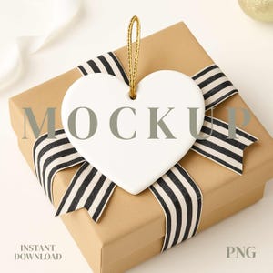 May include: A white heart-shaped ornament with a gold string hangs over a brown gift box. The box is wrapped with a black and white striped ribbon. The word "MOCKUP" is printed on the ornament. The words "INSTANT DOWNLOAD" and "PNG" are also visible.