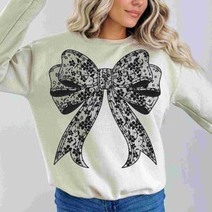 May include: A light beige sweatshirt with a large black lace bow design on the front.