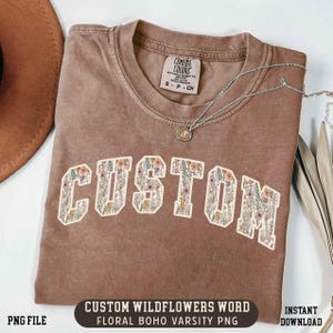 May include: A brown Comfort Colors t-shirt with the word "CUSTOM" in a floral varsity font. The shirt has a crew neck and short sleeves. A silver necklace with a pendant is on the shirt. The text "CUSTOM WILDFLOWERS WORD" is at the bottom.