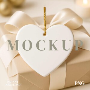 May include: A white heart-shaped ornament with a gold string hangs over a wrapped gift box. The box is tied with a cream-colored ribbon. The word "MOCKUP" is printed on the ornament. The words "INSTANT DOWNLOAD" and "PNG" are also visible.