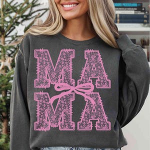 May include: Dark gray sweatshirt with the word "MAMA" in large, pink, floral-patterned letters. A pink bow connects the two "MA" letters. The sweatshirt has a crew neck and long sleeves.