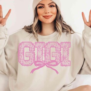 May include: A beige sweatshirt with the name "GIGI" in large, pink, floral-patterned letters. A pink bow is beneath the name. The sweatshirt is a casual style, suitable for everyday wear.