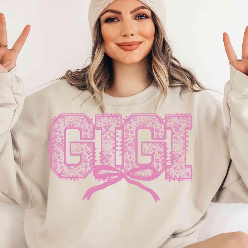 May include: A beige sweatshirt with the name "GIGI" in large, pink, floral-patterned letters. A pink bow is beneath the name. The sweatshirt is a casual style, suitable for everyday wear.