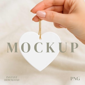 May include: A white heart-shaped ornament with a gold string, held by a hand with manicured nails. The word "MOCKUP" is printed on the ornament in a gray font. The words "INSTANT DOWNLOAD" and "PNG" are also visible.
