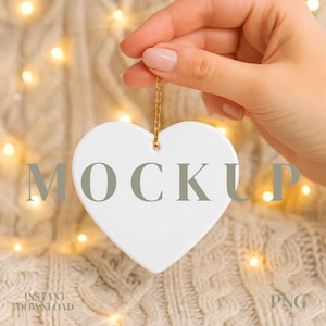 May include: A white heart-shaped ornament with a gold chain, held by a hand. The word "MOCKUP" is printed on the ornament. The background is a knitted fabric with soft, warm lighting. The words "INSTANT DOWNLOAD" and "PNG" are also visible.