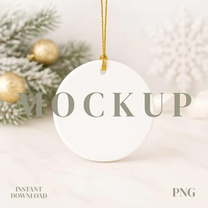 May include: A white ceramic round ornament with a gold string for hanging. The word "MOCKUP" is printed on the ornament. The background includes a Christmas tree branch, gold ornaments, and a snowflake.