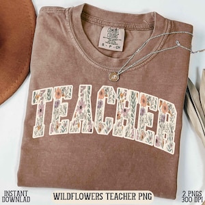 Puede incluir: Una camiseta marrón con la palabra "TEACHER" en un diseño floral. La camiseta tiene cuello redondo y mangas cortas. El texto "WILDFLOWERS TEACHER PNG" está en la parte inferior de la imagen. Un collar plateado con un colgante está sobre la camiseta.