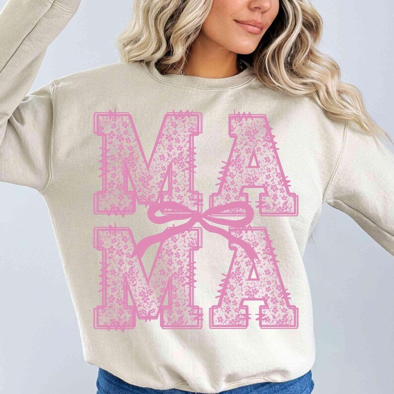 May include: A beige sweatshirt with the word "MAMA" in large, pink, floral-patterned letters. A pink bow is tied between the letters. The sweatshirt is a crew neck style.