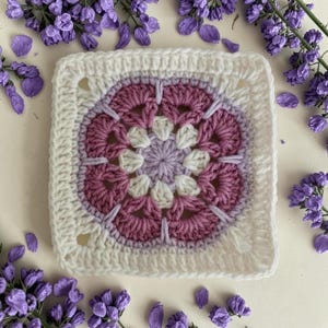 May include: A square, handmade crochet piece with a floral design. The centre is a light purple, surrounded by white, dark pink, and white petals. The outer border is white, with a textured stitch. The piece is surrounded by purple flowers and petals.