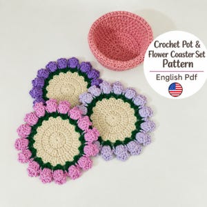 Pode incluir: Um padrão de crochê para um conjunto de porta-copos de flores e uma tigela em formato PDF em inglês. A imagem mostra uma tigela de crochê rosa e três porta-copos circulares com centros creme, folhas verdes e pétalas roxas ou rosas ao redor.