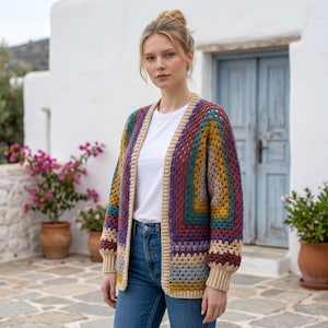 Op de afbeelding: Een gehaakt vest met lange mouwen en een veelkleurig granny square-patroon in tinten paars, mosterdgeel, teal en bordeauxrood, afgewerkt met een effen beige geribbelde boord langs de opening aan de voorzijde, de mouwen en de onderkant.