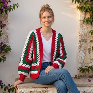 May include: A crochet cardigan featuring a granny square pattern in alternating stripes of red, white, and green. The garment has long sleeves with ribbed red cuffs, a V-neckline, and a solid red ribbed trim along the front opening and hem.