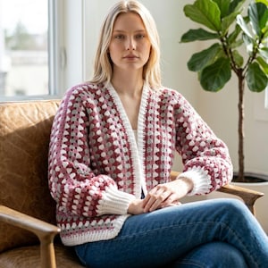 May include: A handmade, open-front crochet cardigan in a granny square pattern. The sweater features a color scheme of burgundy, white, and gray. The sleeves and trim are white, and the garment is worn by a person sitting in a chair.