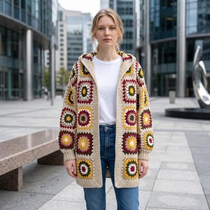 May include: A hooded cardigan constructed from multicolored granny squares in shades of cream, maroon, olive green, and yellow. The garment features long sleeves, a front-open design, and ribbed cuffs, worn over a plain white t-shirt and blue jeans.