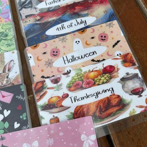 May include: A collection of decorative budget envelopes for various holidays, including Father's Day, 4th of July, Halloween, Thanksgiving, and Christmas. Each envelope features themed illustrations and text labels.