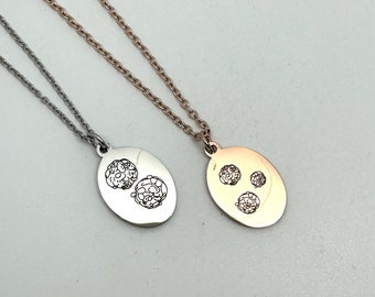 May include: Two silver necklaces with oval pendants. One pendant has a design of three small circles, the other has a design of two larger circles. The necklace on the left is silver, the necklace on the right is rose gold.
