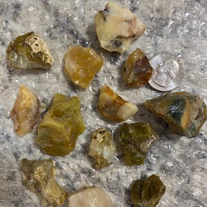 May include: A collection of raw, uncut gemstones in shades of yellow, green, and brown. The stones vary in size and shape, with a quarter coin included for size reference. The gemstones are displayed on a textured, clear surface.