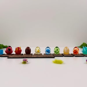 Könnte beinhalten: Eine Sammlung farbenfroher, dekorativer Eier, jedes mit einzigartigen Designs und Farben, präsentiert auf kleinen Holzplattformen. Die Eier variieren in der Farbe, darunter Rot, Gelb, Blau und Grün. Die Szene ist mit Moos und kleinen floralen Akzenten dekoriert.