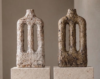 May include: Two tall, rectangular ceramic vases with a rough, textured surface. One vase is off-white with brown speckles, and the other is a dark, mottled brown. Both feature a central vertical cutout and a narrow neck opening at the top.
