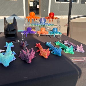 May include: A collection of 3D printed, colorful, small, dragon-like figures. The figures are arranged in a row on a black surface. The figures are painted in a rainbow of colors, from blue to red.