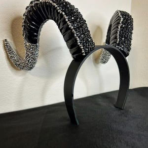May include: A black headband featuring curved ram horns. The horns are black with silver crystal detailing. The headband is made of black material and is designed to be worn on the head.
