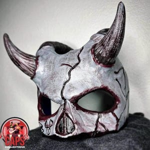 May include: A white, textured mask featuring two prominent, dark-tipped horns and deep red, vein-like cracks across the surface. The mask has two large eye openings and a central nose cavity, with dark shading around the edges and crevices.