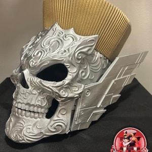 Puede incluir: Un casco con forma de calavera plateada que presenta intrincados patrones de volutas en relieve, una cresta alta plisada de color dorado en la parte superior y aletas laterales plateadas angulares. Un logo circular muestra el texto CAPS COLLECTIONS.