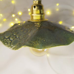 Puede incluir: Lámpara colgante artesanal con pantalla de cerámica verde texturizada. La pantalla está unida a un portalámparas color latón y un cordón de cuerda natural. La bombilla es visible debajo de la pantalla. Luces de hadas amarillas en el fondo.